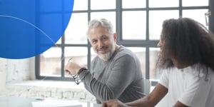 Older man sitting at a table in a bight room holding his glasses smiling at the camera