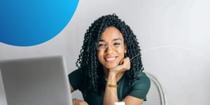 Woman sitting at desk with a laptop with hand to her chin and smiling in camera 
