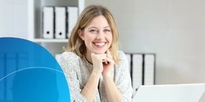 Woman sitting at desk with hands under her chin smiling at camera