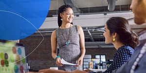Woman standing and smiling at other female employees holding paper in hand 