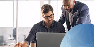 man wearing glasses sitting down and looking into laptop and another man standing next to him