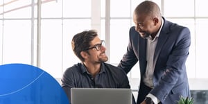 Male employee sitting at desk with laptop looking up at another male employee and smiling