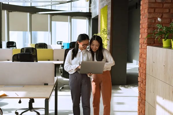 Two female employees standing together in office looking into laptop screen